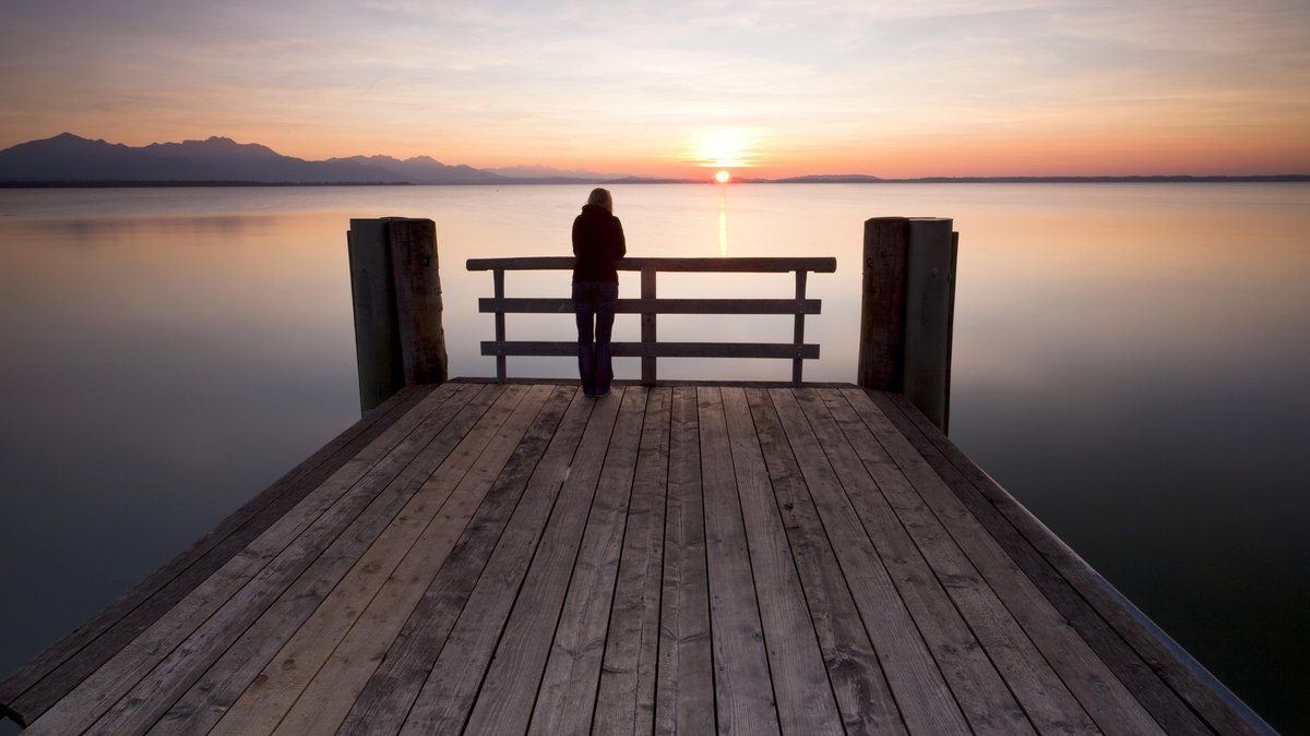 Frau steht alleine an einem Steg und betrachtet den Sonnenuntergang