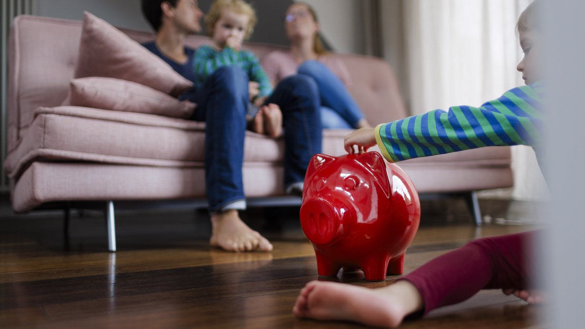 Eine Familie mit zwei Kindern sitzt im Wohnzimmer, ein rotes Sparschwein steht auf dem Boden.