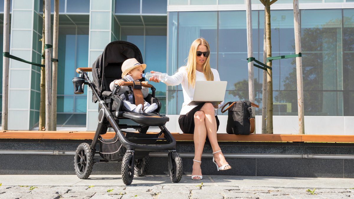 Eine Mutter im Businesslook arbeitet mit Laptop auf dem Schoß auf einer Treppe und hält gleichzeitig ihrem Kleinkind im Kinderwagen eine Wasserflasche hin.