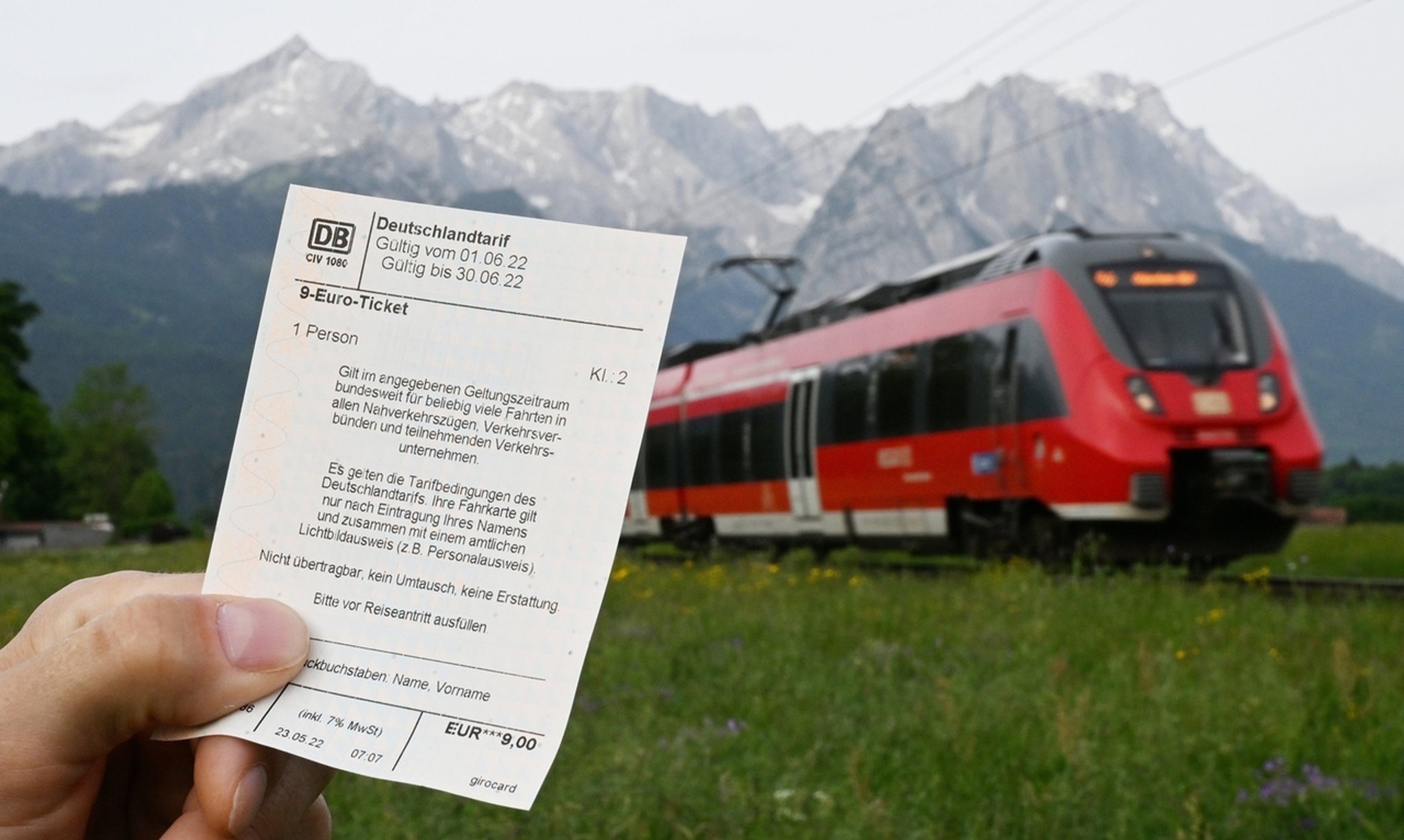 ILLUSTRATION - 23.05.2022, Bayern, Garmisch-Partenkirchen: Ein Mann hält ein 9-Euro-Ticket in der Hand, im Hintergrund fährt ein Zug der Deutschen Bahn vor der Bergkulisse des Wettersteingebirges vorbei (gestelltes Foto). Die Bundesregierung hat Ende März 2022 ein Energie-Entlastungspaket vorgestellt. Darin ist unter anderem ein zeitlich begrenztes 9-Euro-Ticket für den öffentlichen Personennahverkehr vorgesehen. Foto: Angelika Warmuth/dpa +++ dpa-Bildfunk +++