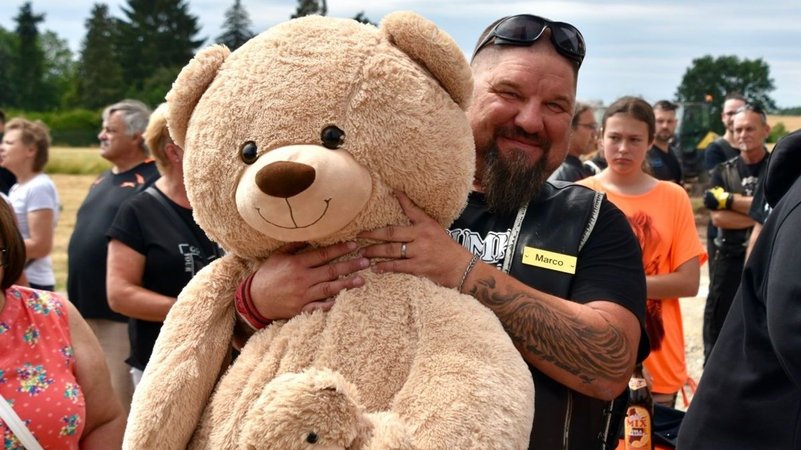 Ein Biker hält einen großen Teddybären im Arm. Im Hintergrund sind weitere Menschen zu sehen.