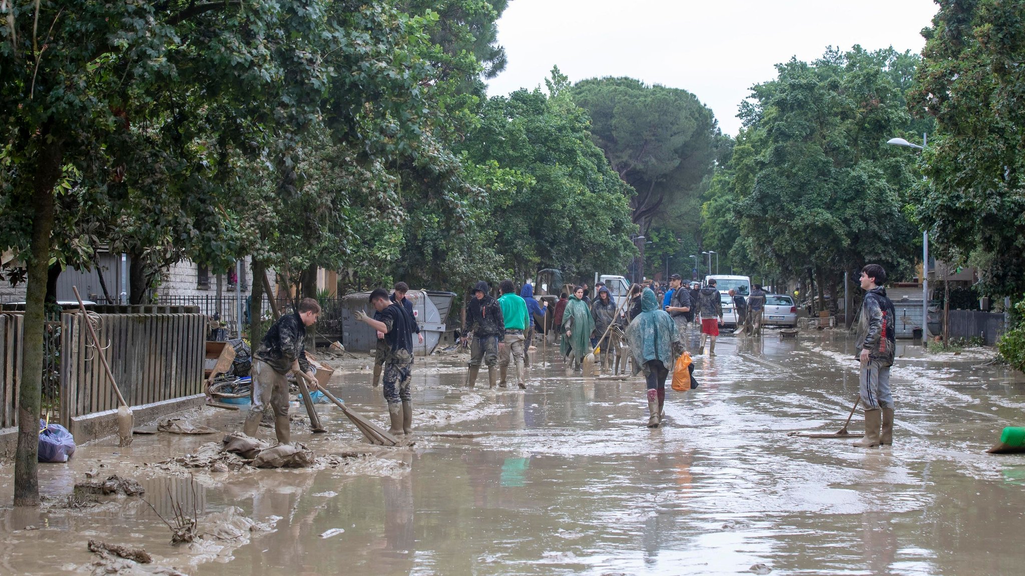 Eine von Schlamm und Wasser überzogene Straße in Faenza in Italien. Menschen in Regenkleidung und Arbeitsgeräten sind mit Aufräumarbeiten beschäftigt.