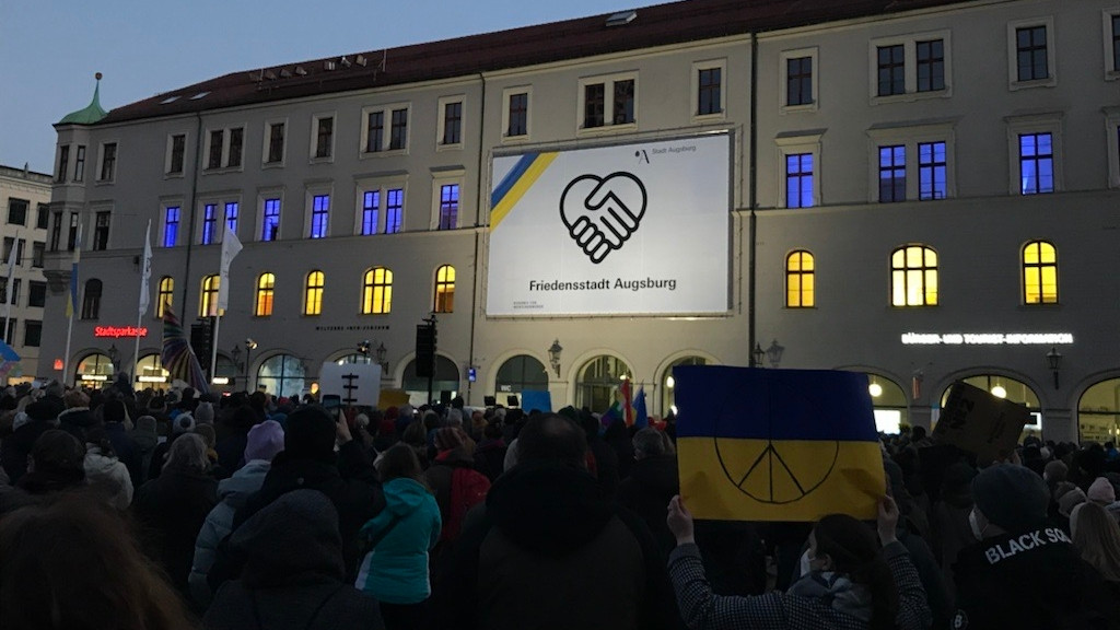 Viele Menschen stehen auf dem Rathausplatz in Augsburg vor einem Plakat auf dem steht: Friedensstadt AugsburgDie KundAuf dem 