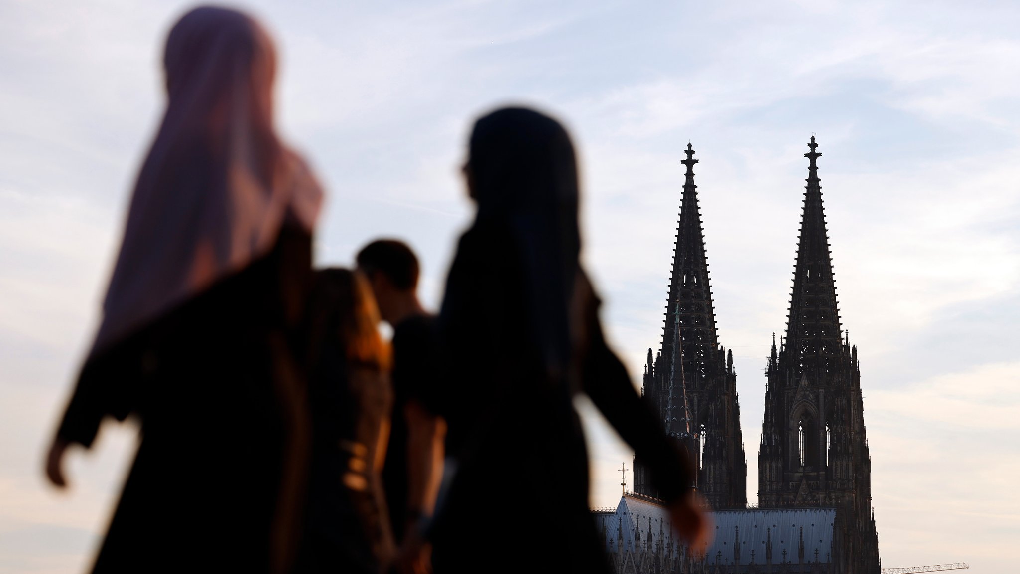 Frauen mit Kopftuch am Kölner Rheinboulevard, im Hintergrund ragt die Doppelspitze des Kölner Doms in den Himmel. 