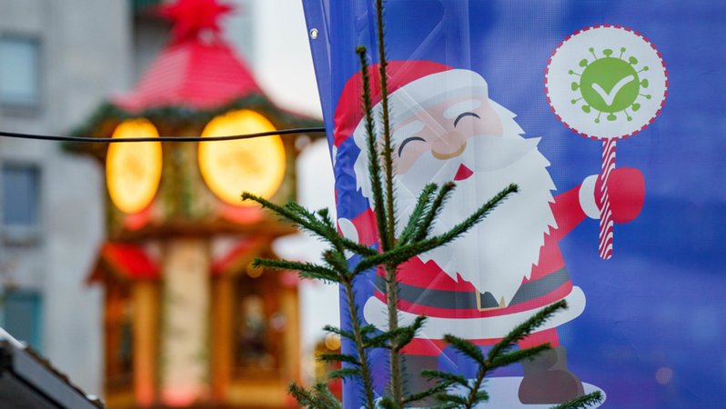 Ein Weihnachtsmann mit einem stilisierten Impfnachweis ist auf einem Plakat am Eingang eines Weihnachtsmarktes zu sehen.