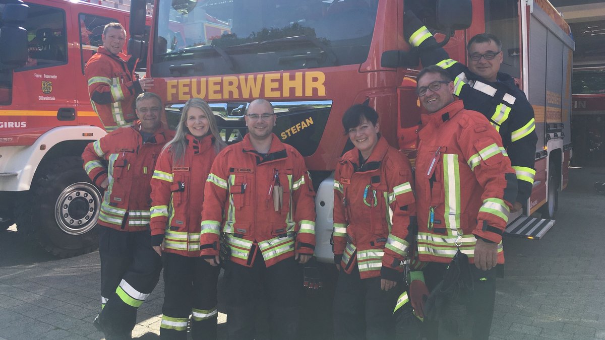 Sieben Frauen und Männer in Schutzkleidung stehen vor einem Feuerwehrauto. 