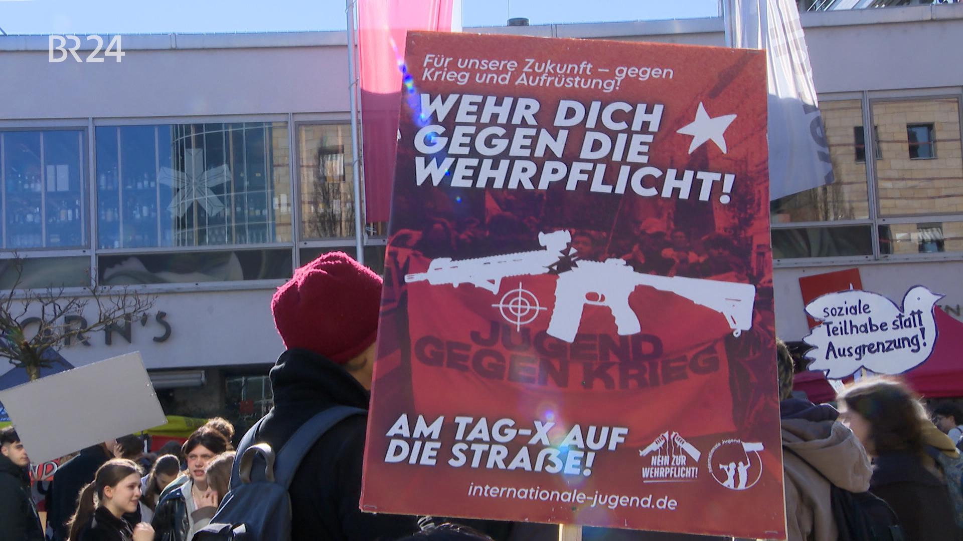 Im Vordergrund ein rotes Plakat mit der Aufschrift "WEHR DICH GEGEN WEHRPFLICHT! AM TAG-X AUF DIE MAXSTRASSE". Im Hintergrund ist dein Demonstrationszug zu sehen. 