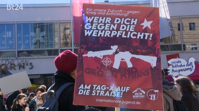 Im Vordergrund ein rotes Plakat mit der Aufschrift "WEHR DICH GEGEN WEHRPFLICHT! AM TAG-X AUF DIE MAXSTRASSE". Im Hintergrund ist dein Demonstrationszug zu sehen. | Bild: Bayerischer Rundfunk 2026 Im Vordergrund ein rotes Plakat mit der Aufschrift "WEHR DICH GEGEN WEHRPFLICHT! AM TAG-X AUF DIE MAXSTRASSE". Im Hintergrund ist dein Demonstrationszug zu sehen.
