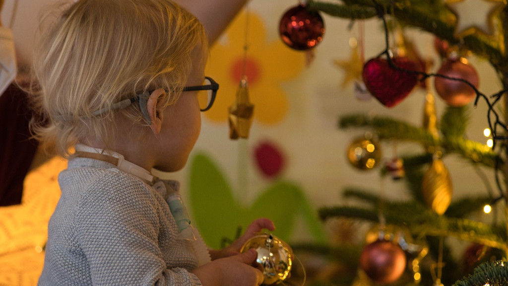 Zu sehen ist ein Mädchen mit einem Beatmungsschlauch und einer Christbaumkugel in der Hand vor einem Weihnachtsbaum