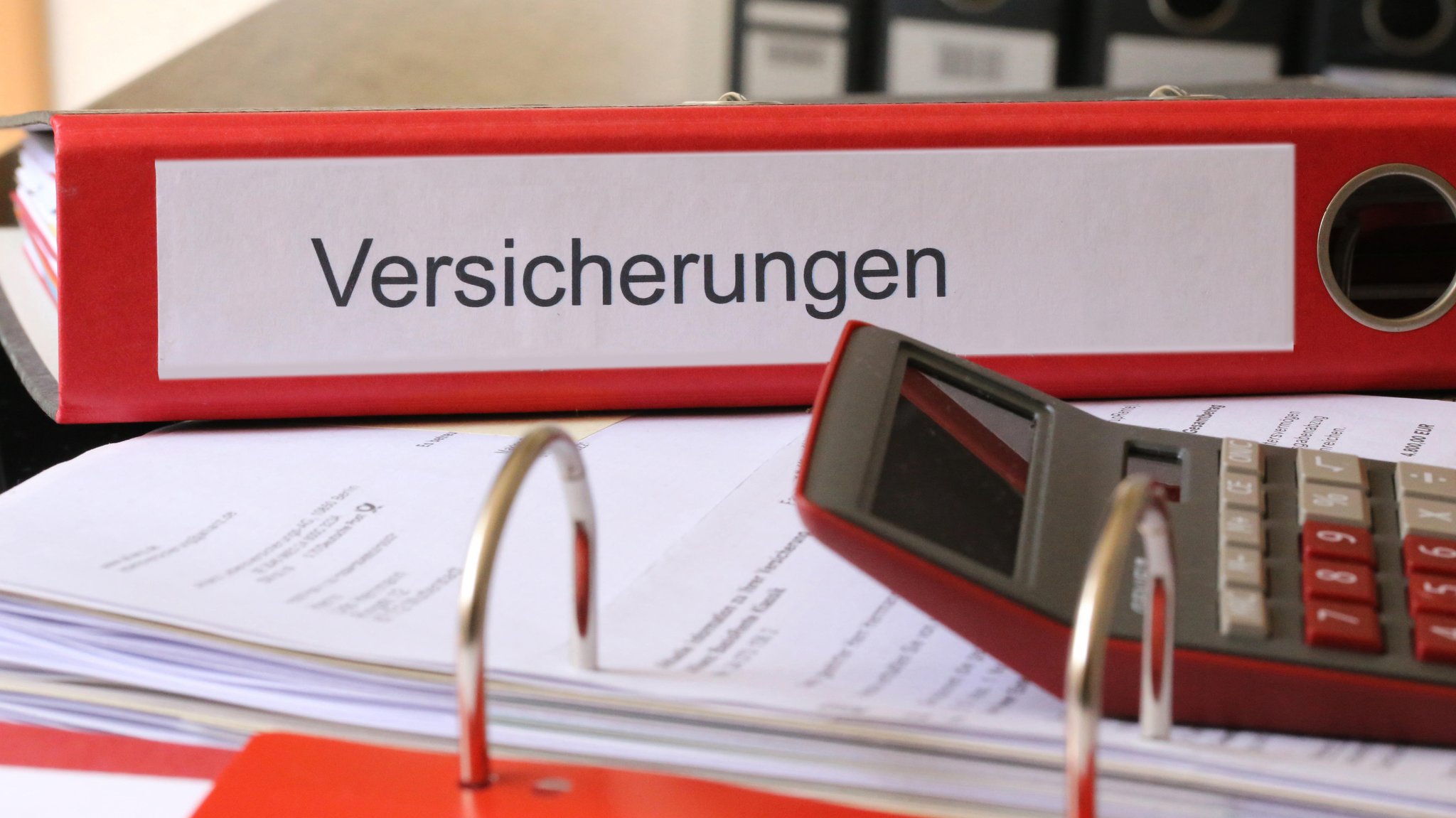 Ein roter Ordner mit der Aufschrift "Versicherungen" liegt auf einem Tisch.