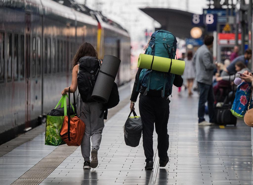 Junge Reisende gehen in einem Hauptbahnhof mit ihren Rucksäcken über den Bahnsteig.