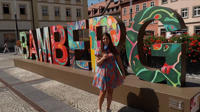 BR-Korrespondentin Sandra Jozipovic steht auf dem Bamberger Maxplatz, dahinter sind großen Buchstaben der Schriftzug "Bamberg" zu lesen. 