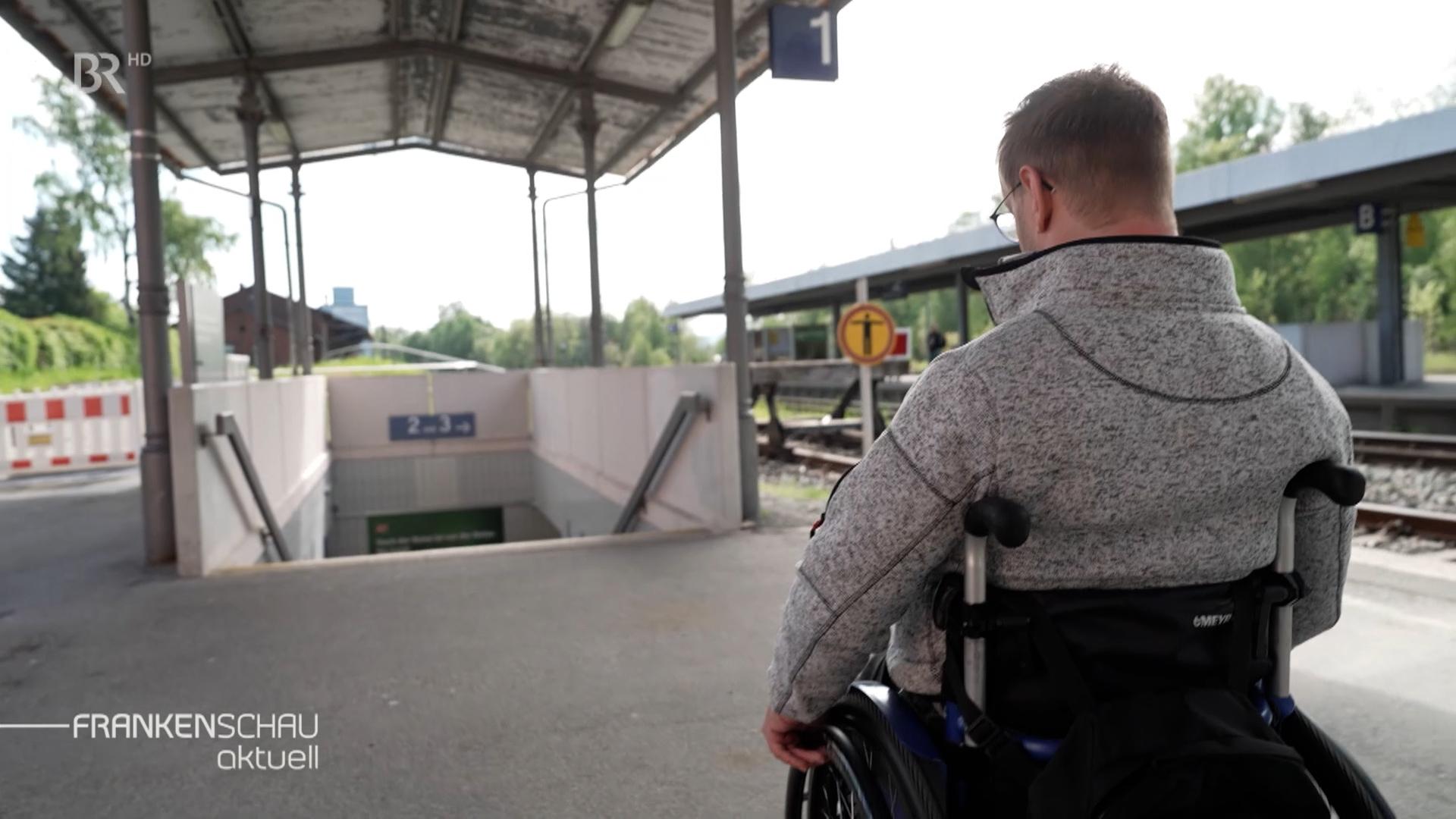 Ein Rollstuhlfahrer steht an einem Bahnhof mit seinem Rollstuhl vor Treppenstufen. 