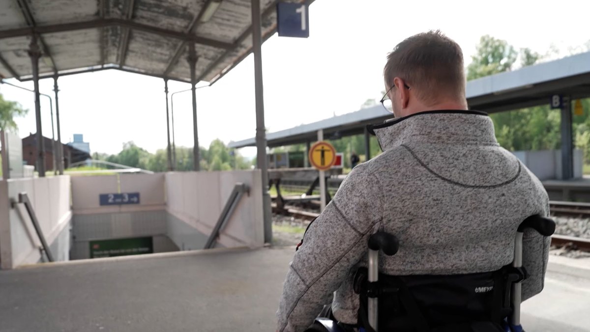 Ein Rollstuhlfahrer steht an einem Bahnhof mit seinem Rollstuhl vor Treppenstufen. 