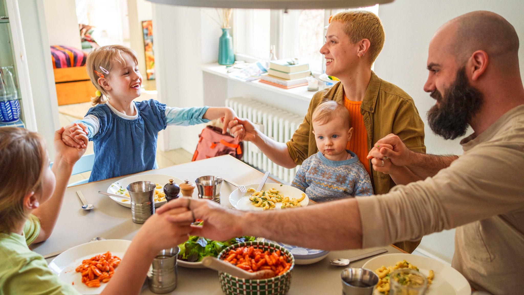 Ein Mann und eine Frau sitzen mit drei Kindern um einen gedeckten Tisch und halten sich an den Händen.