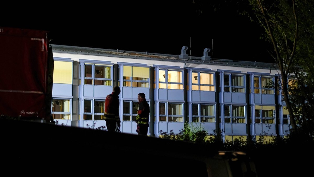 Zwei Feuerwehrmänner stehen nachts vor einem hell beleuchteten Krankenhaus. 