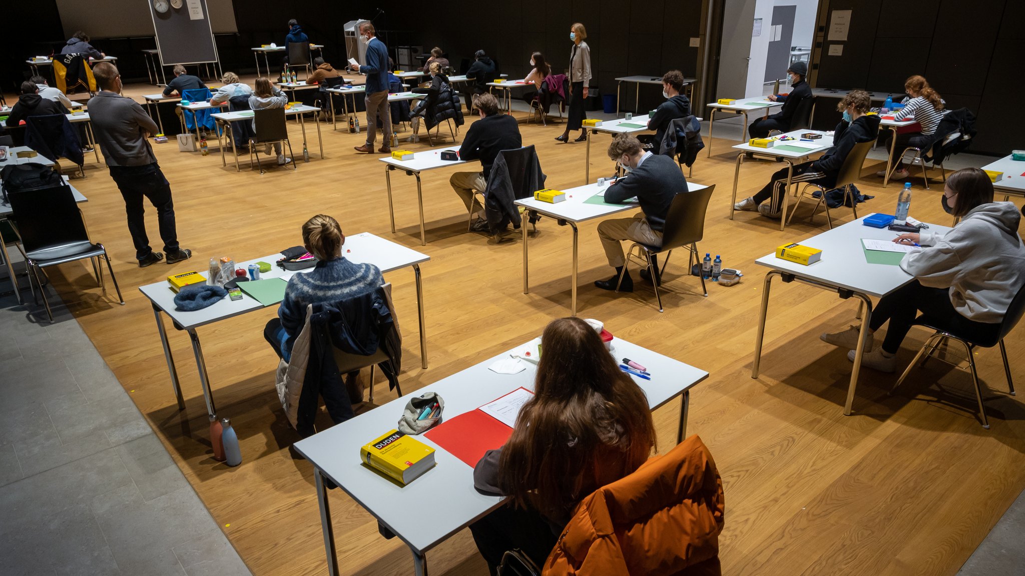 Schüler sitzen unter der Aufsicht mehrerer Lehrer in einer Aula um eine Prüfung abzulegen.