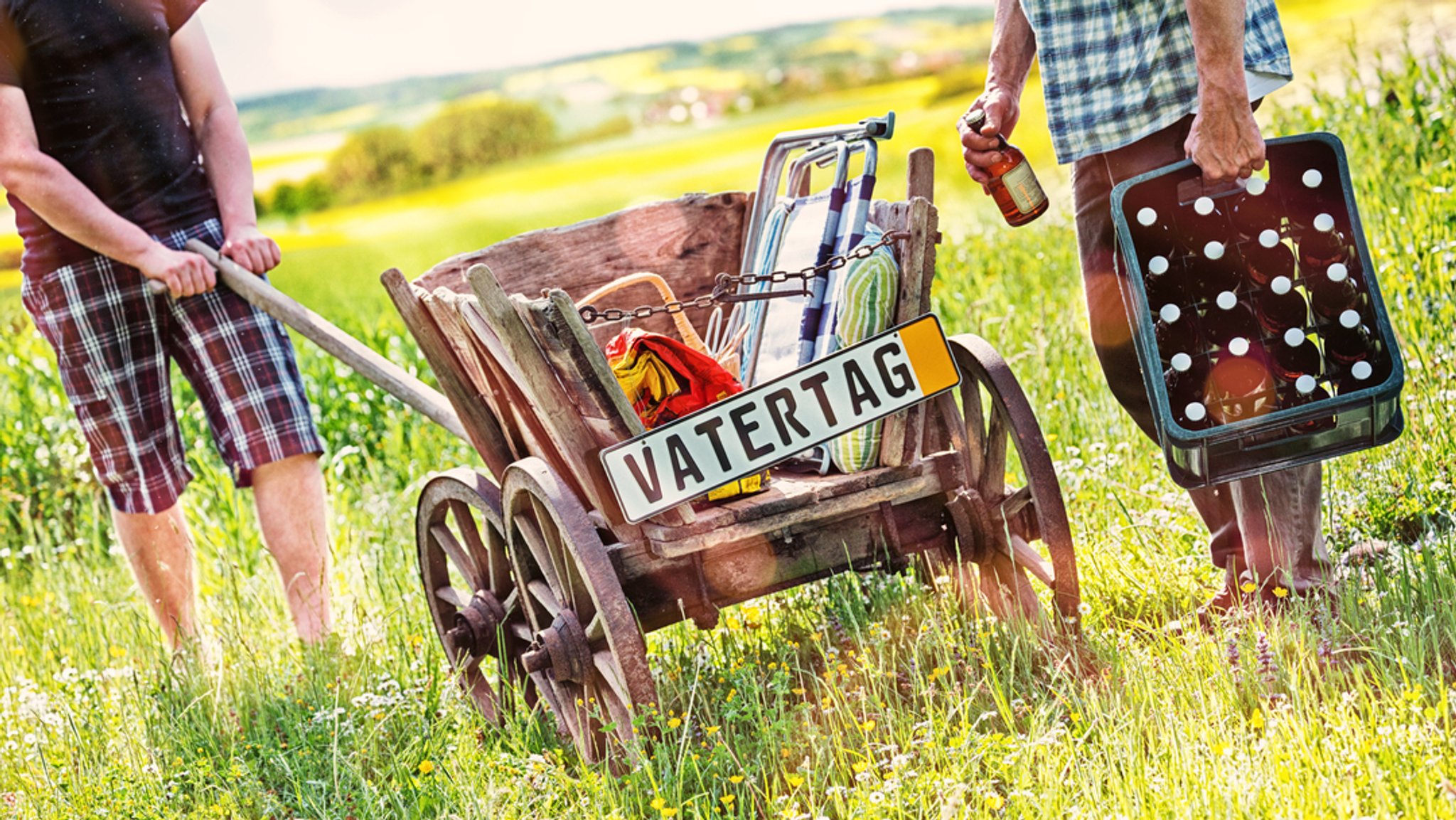 Zwei Männer mit einem Bierkasten und einem Bollerwagen mit der Aufschrift "Vatertag".