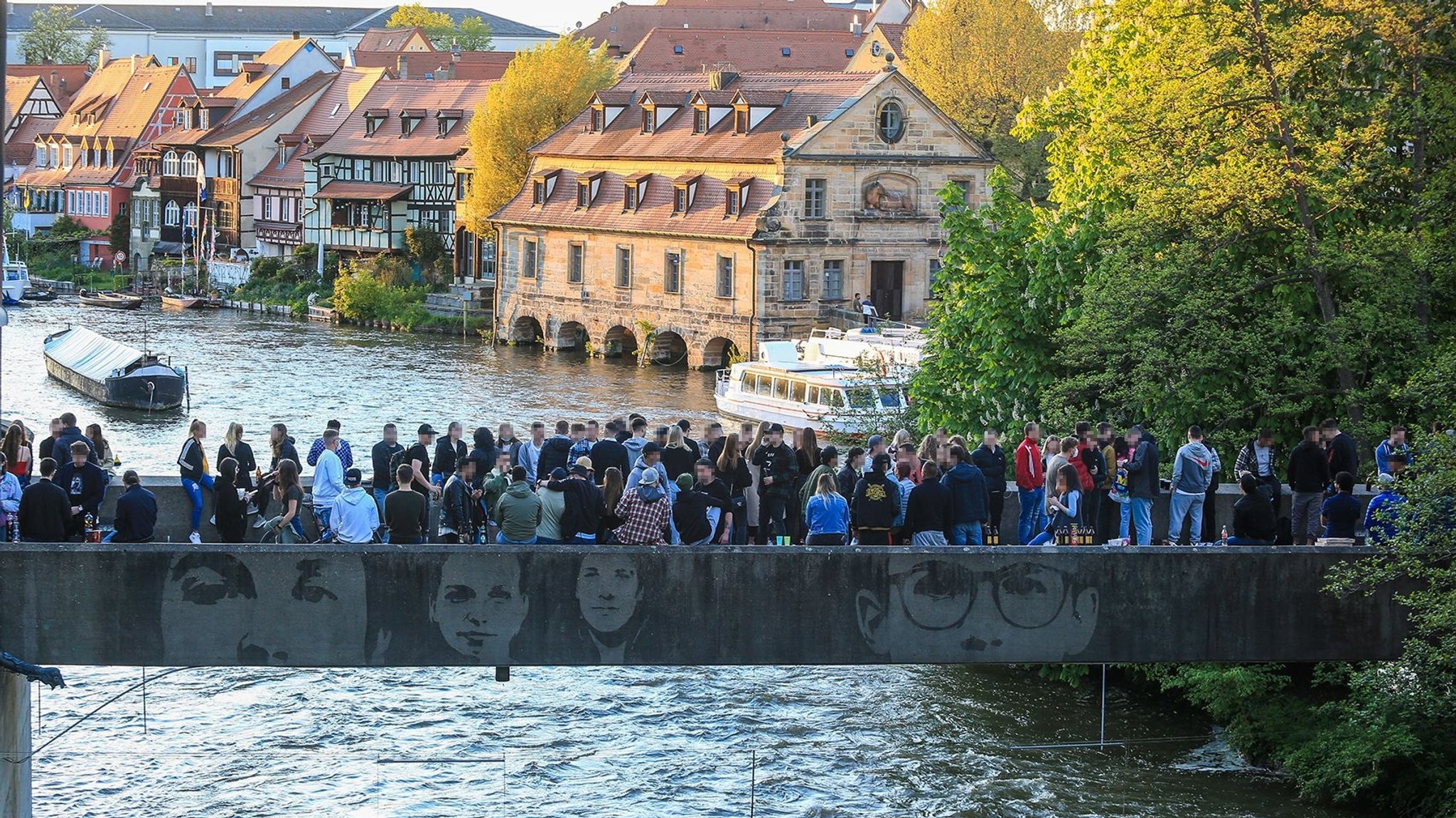 Auf einer Brücke stehen viele junge Leute, im Hintergrund Schiffe auf dem Wasser.