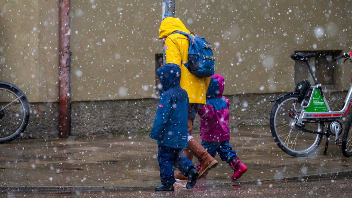 Eine Frau geht mit zwei Kindern bei Schneefall über eine Straße. In München hat Mitte April nochmal kurz der Winter vorbeigeschaut. 