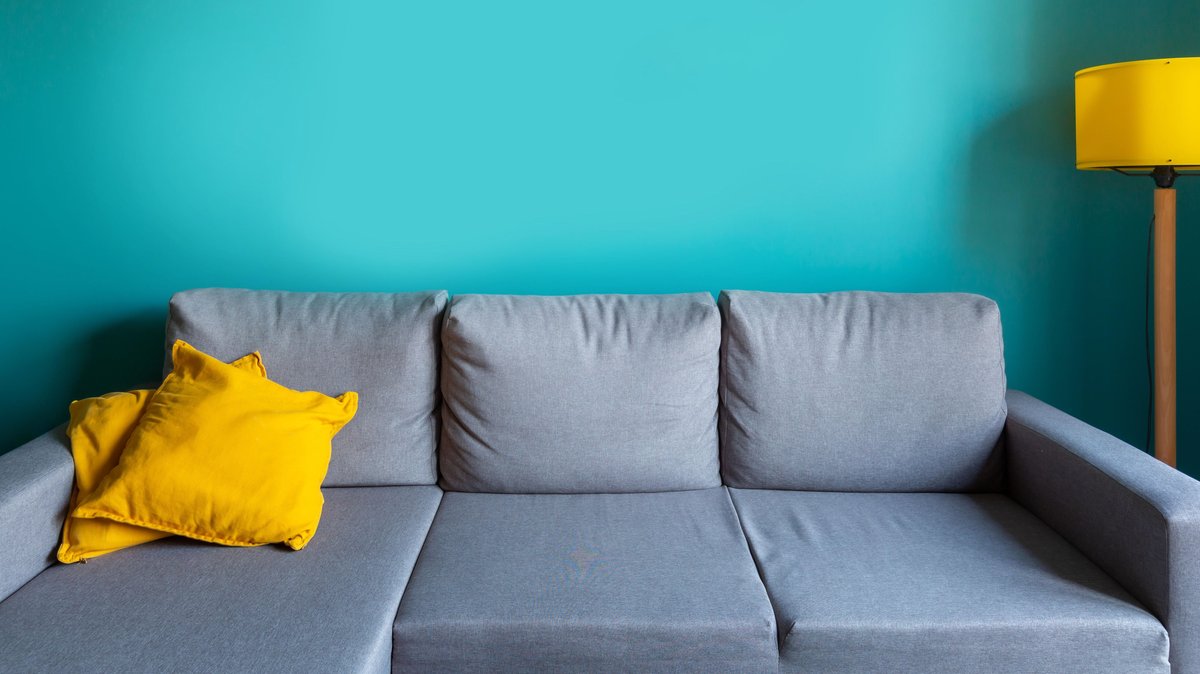 Ein graues Sofa mit gelben Kissen und gelber Lampe vor einer türkisfarbenen Wand (Symbolbild)