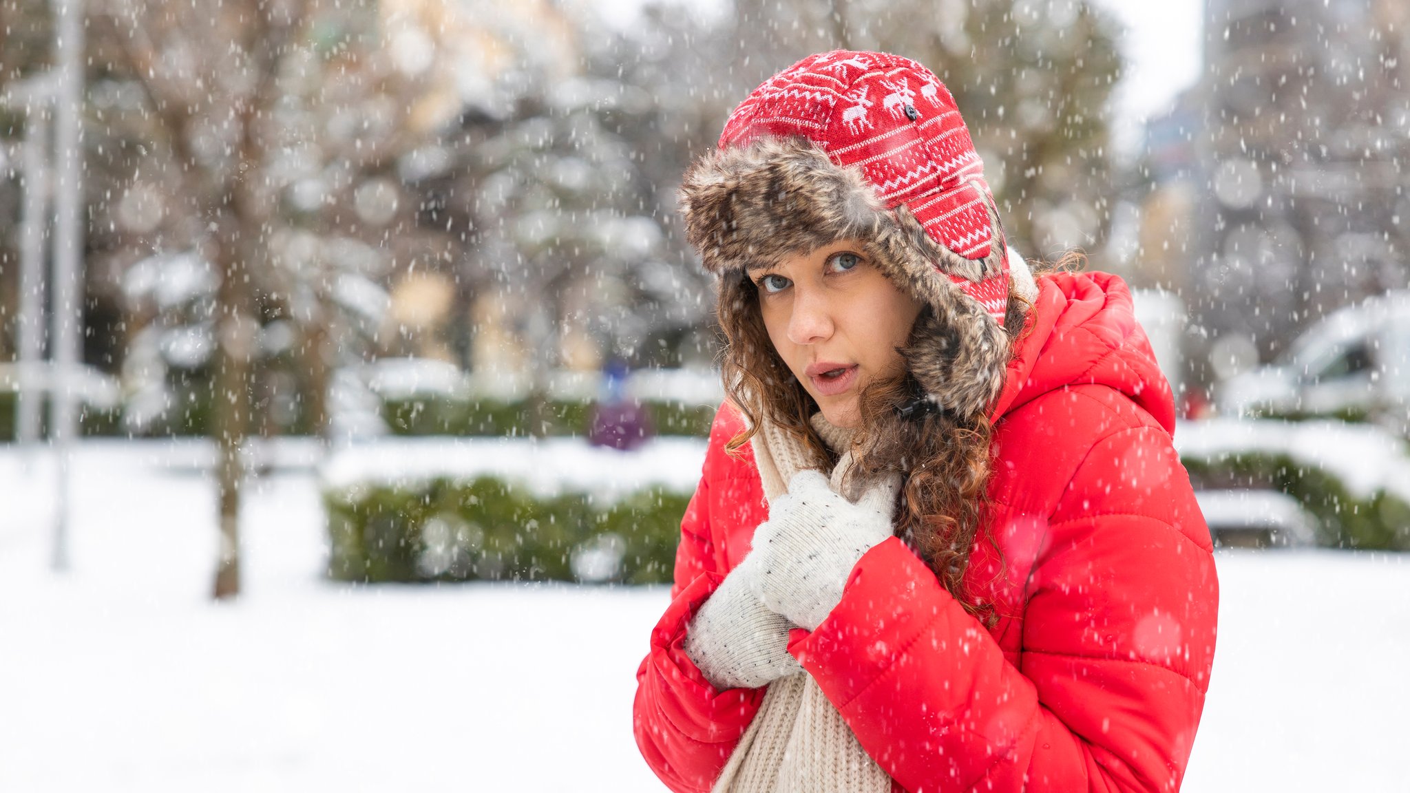 Frau mit rotem Anorak und Mütze steht frierend im Schnee