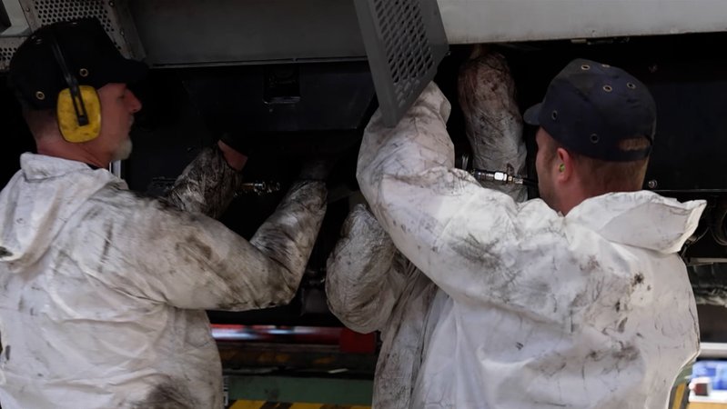 Zwei Männer in weißen Arbeitsanzügen führen unter einem Fahrzeug Reparaturarbeiten durch.