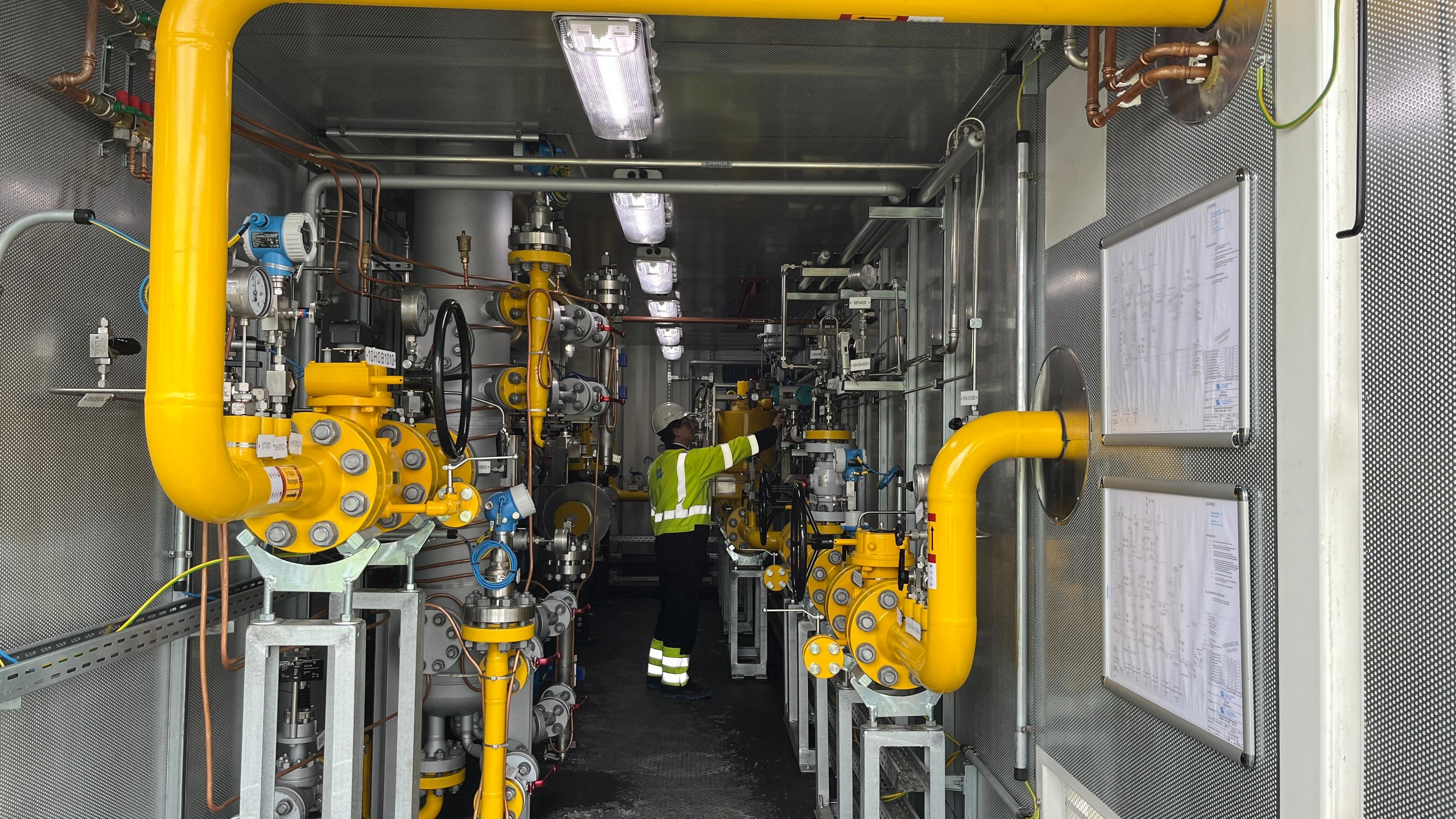 Ein Mann in Schutzkleidung steht zwischen vielen Rohren, Leitungen und Kabeln.