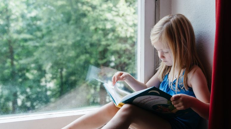 Ein kleines Mädchen sitzt auf der Fensterbank und sieht sich ein Kinderbuch an. | Bild:BR/Sylvia Bentele