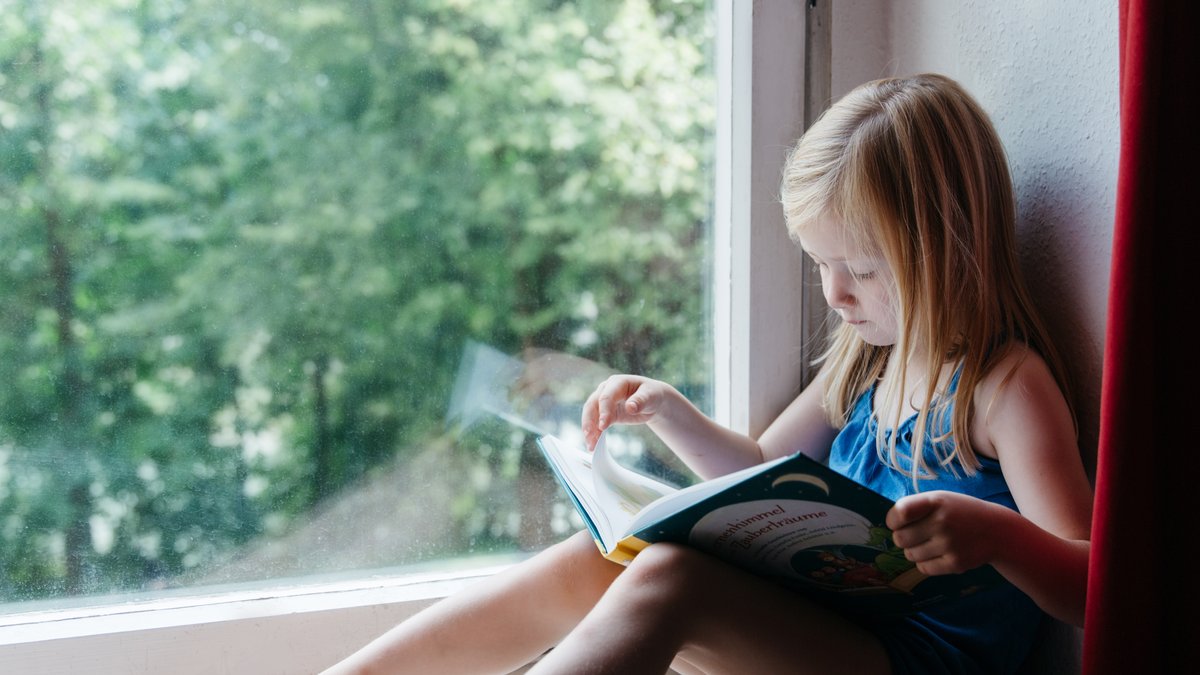 Ein kleines Mädchen sitzt auf der Fensterbank und sieht sich ein Kinderbuch an.