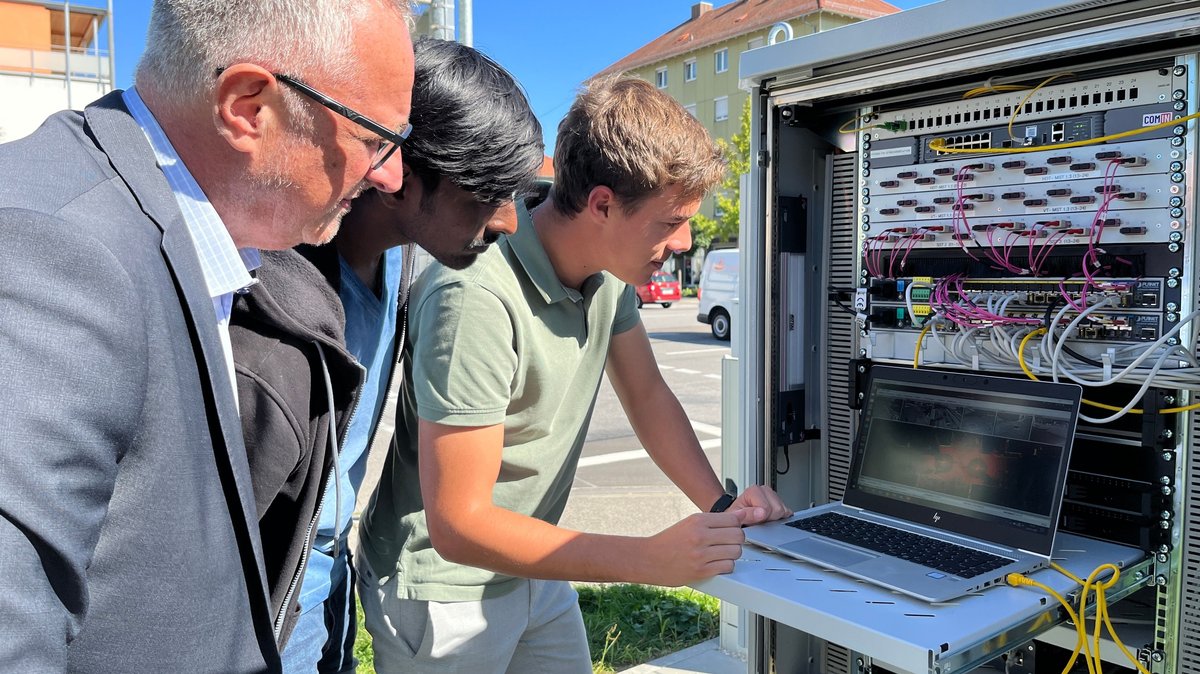 Drei Männer stehen an einer Straßenkreuzung an einem Kasten, aus dem viele Kabel herausragen, an denen ein Laptop angeschlossen ist. Auf dem Bildschirm erkennt man schemenhaft die Bilder der Kameras und Sensoren des Projekts KIVI an der Kreuzung.