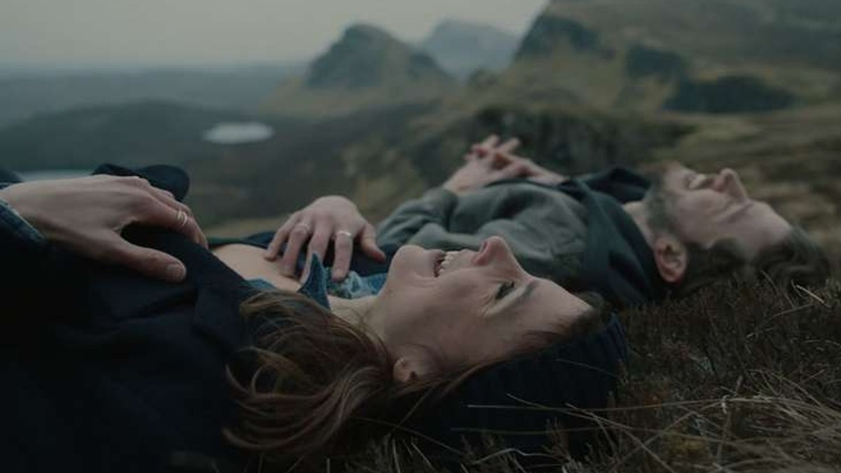 Eine junge Frau und ein junger Mann liegen mit dem Rücken auf einer wolkenverhangenen Wiese  inGebirgslandschaft.