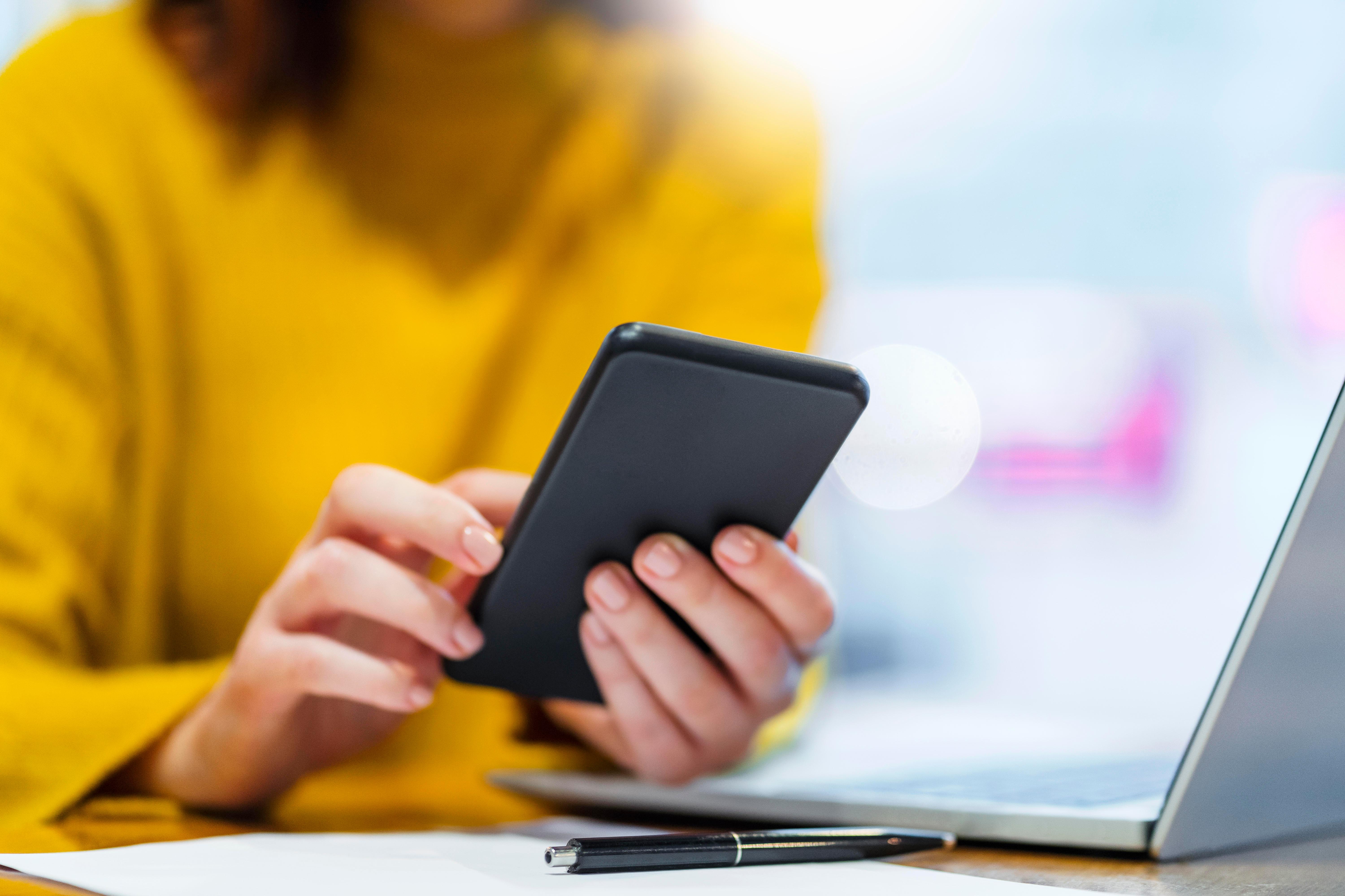 Frau tippt auf ihr Handy, daneben steht ein Laptop