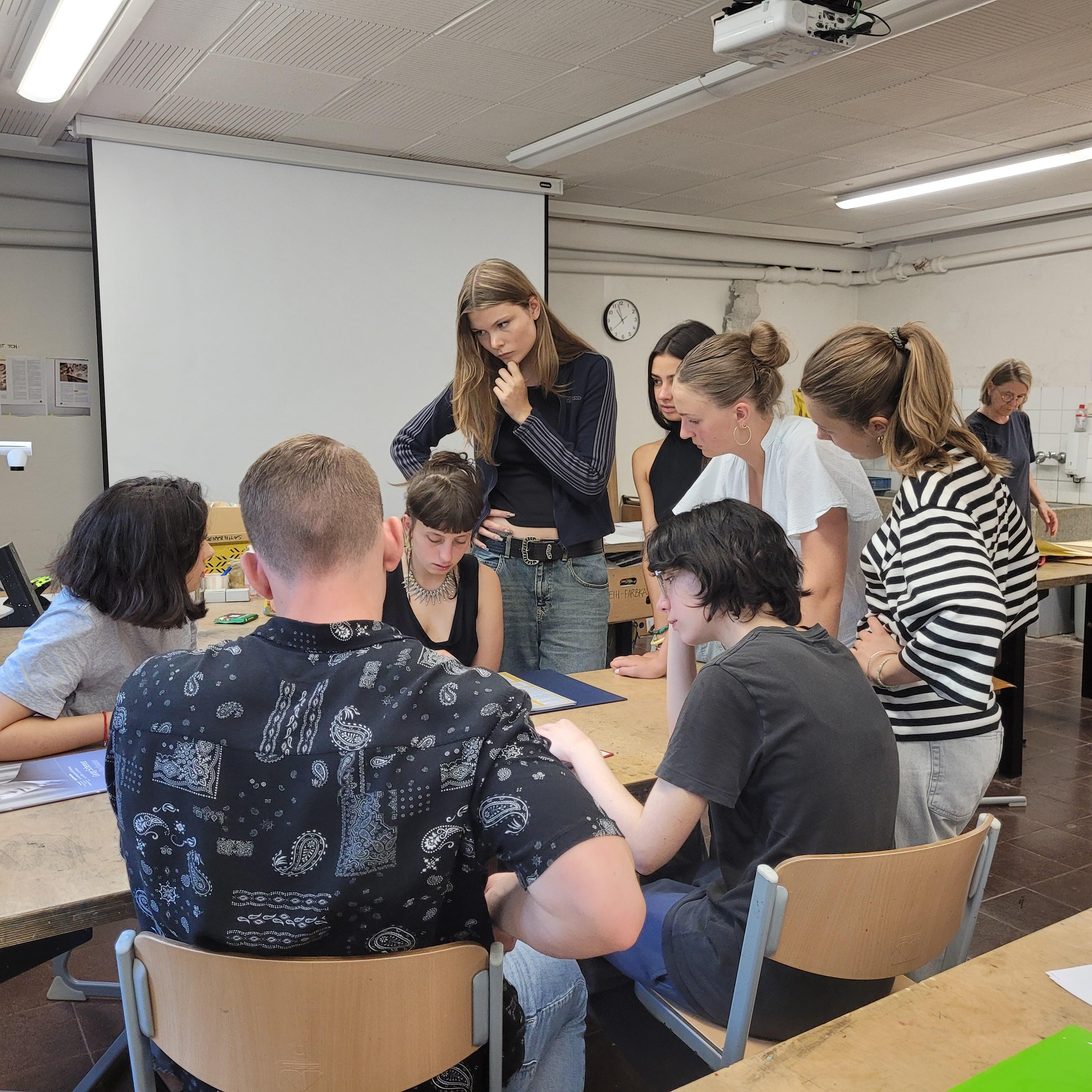 Eine Gruppe von acht Schülerinnen und Schülern steht und sitzt um einen Tisch herum auf dem Rechercherunterlagen aus dem Erinnerungsprojekt liegen.