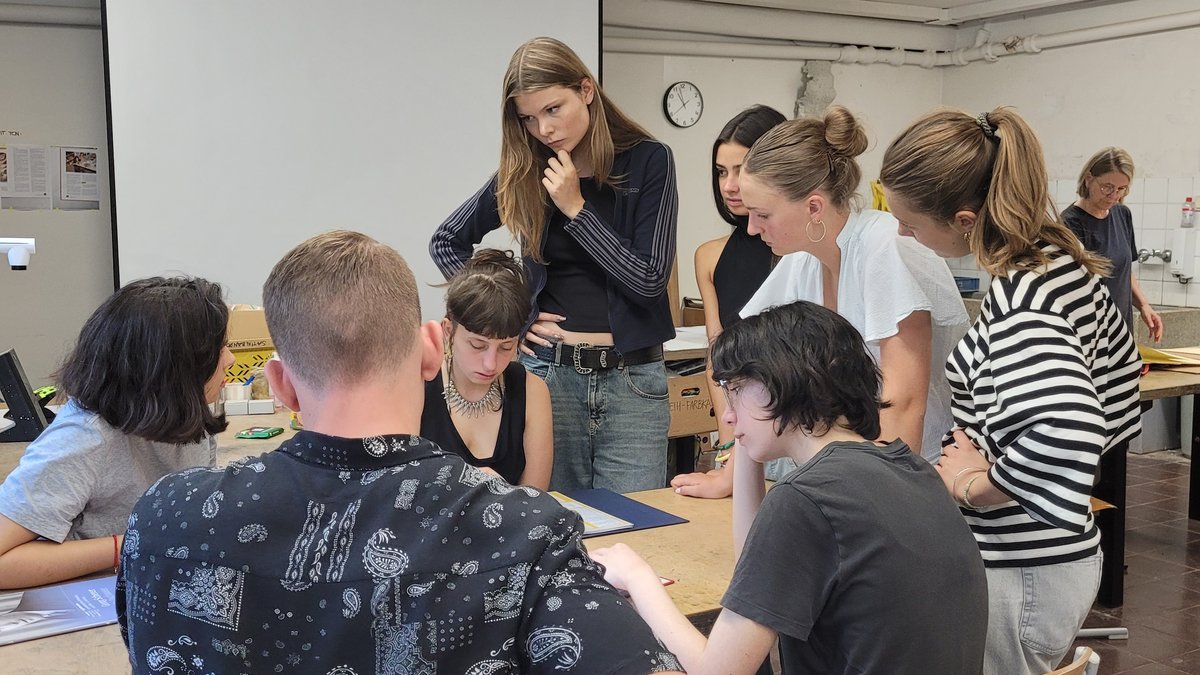 Eine Gruppe von acht Schülerinnen und Schülern steht und sitzt um einen Tisch herum auf dem Rechercherunterlagen aus dem Erinnerungsprojekt liegen.