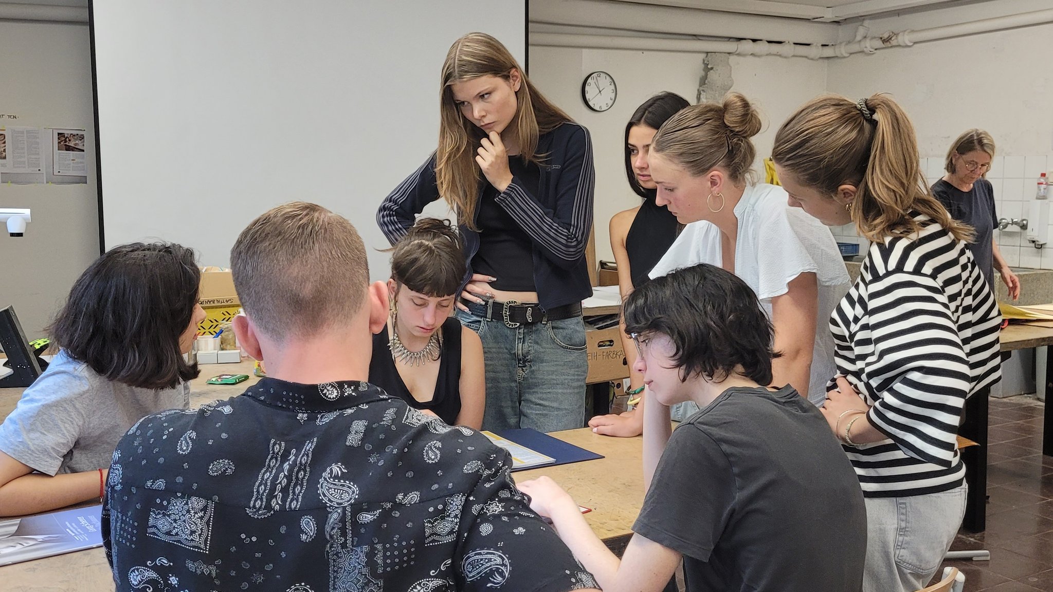 Eine Gruppe von acht Schülerinnen und Schülern steht und sitzt um einen Tisch herum auf dem Rechercherunterlagen aus dem Erinnerungsprojekt liegen.