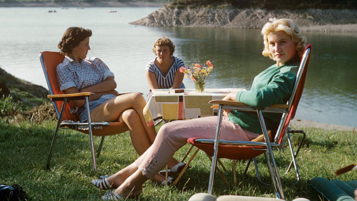 Drei Frauen aus den sechziger Jahren sitzen auf Campingstühlen am Ufer eines Flusses.