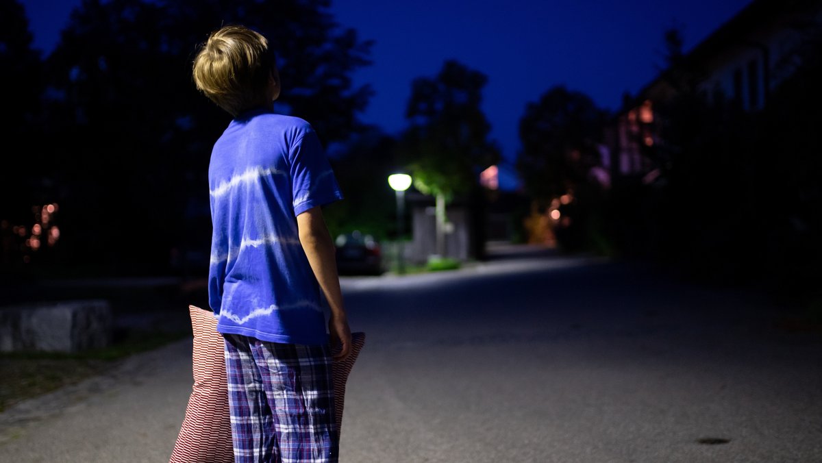 Ein Junge im Schlafanzug steht nachts mit einem Kissen in der Hand auf einer Straße.