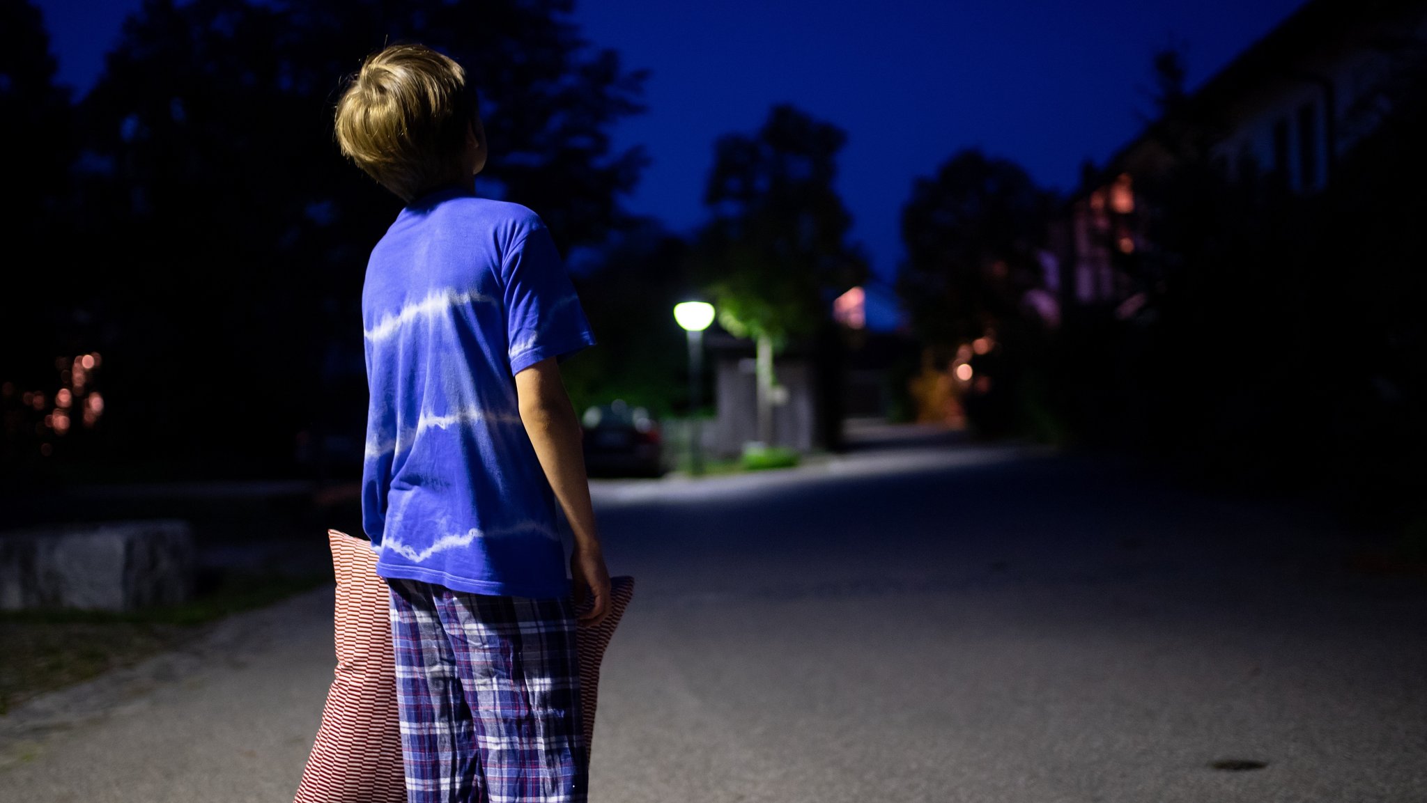 Ein Junge im Schlafanzug steht nachts mit einem Kissen in der Hand auf einer Straße.