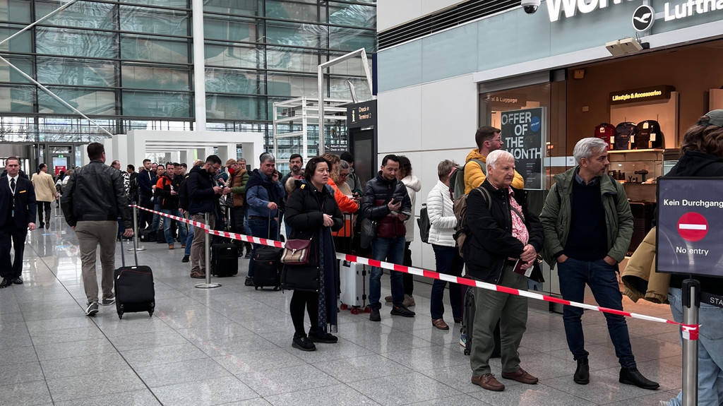 Menschen stehen am Münchner Flughafen in einer langen Warteschlange, die mit Absperrband markiert ist.