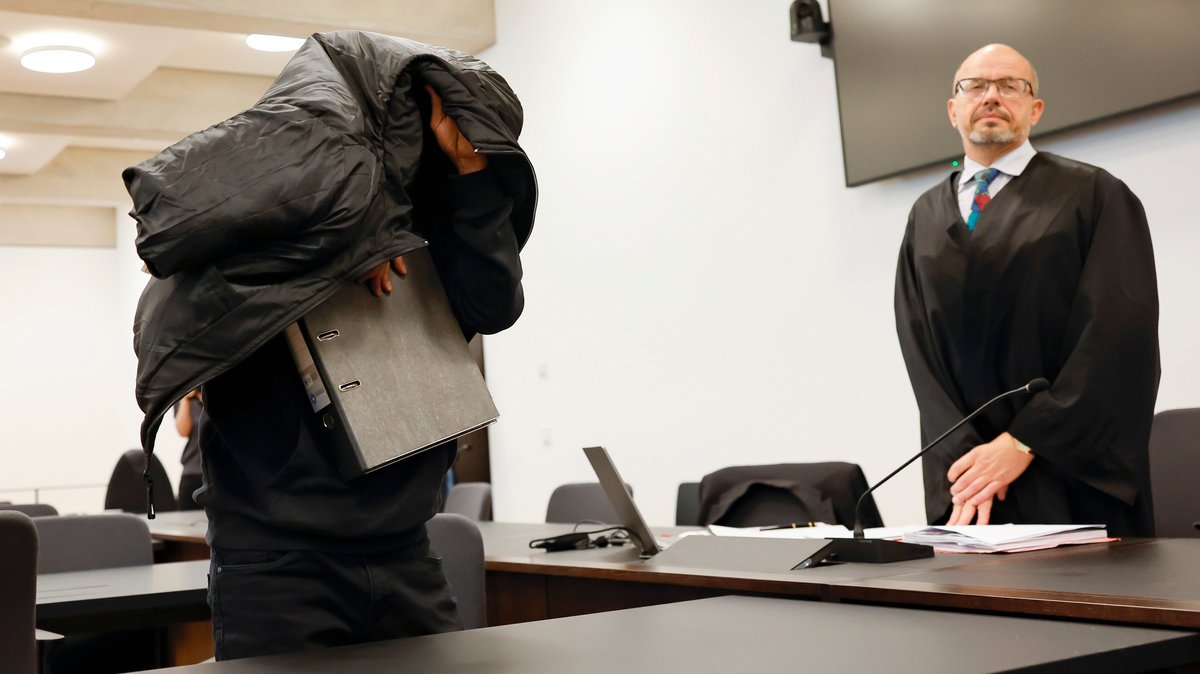 Ein Mann steht im Gerichtssaal und verdeckt seinen Kopf mit einer schwarzen Jacke.