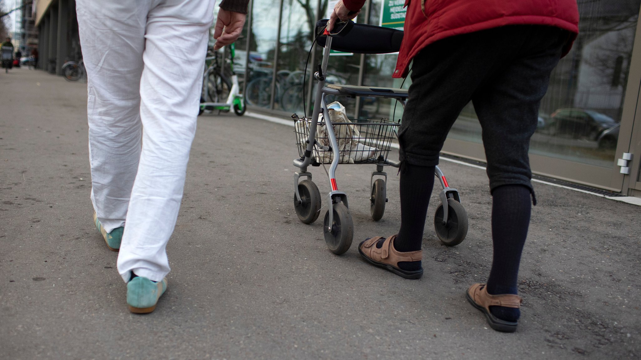 Alter Mann geht mit einem Rollator auf der Straße, begleitet von einem Helfer