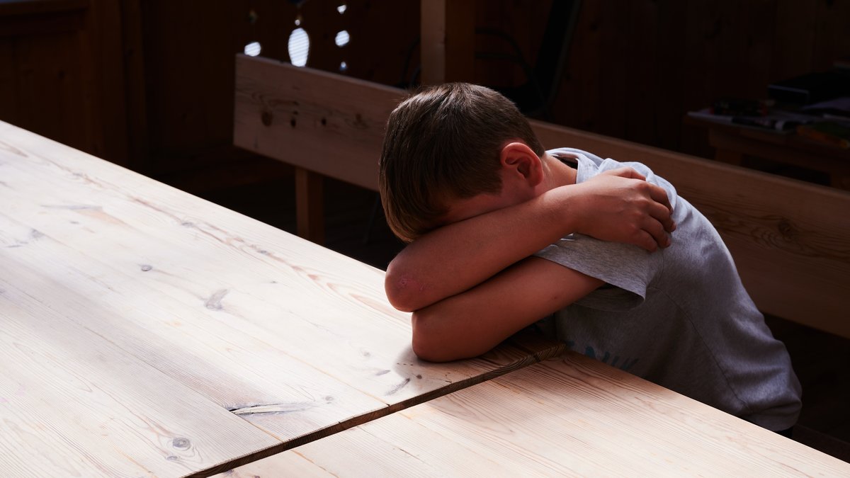Ein Kind versteckt sein Gesicht unter seinen Armen und liegt mit dem Kopf auf einem Tisch. (gestellte Szene)