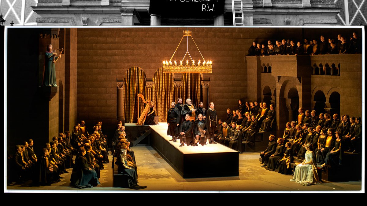 Szene der Oper "Tannhäuser und der Sängerkrieg auf Wartburg": Das Bühnenbild zeigt den Innenraum einer Burg, in der zahlreiche Personen zu einer kleinen Gruppe schauen, die erhöht auf einem beleuchteten Podest steht. 
