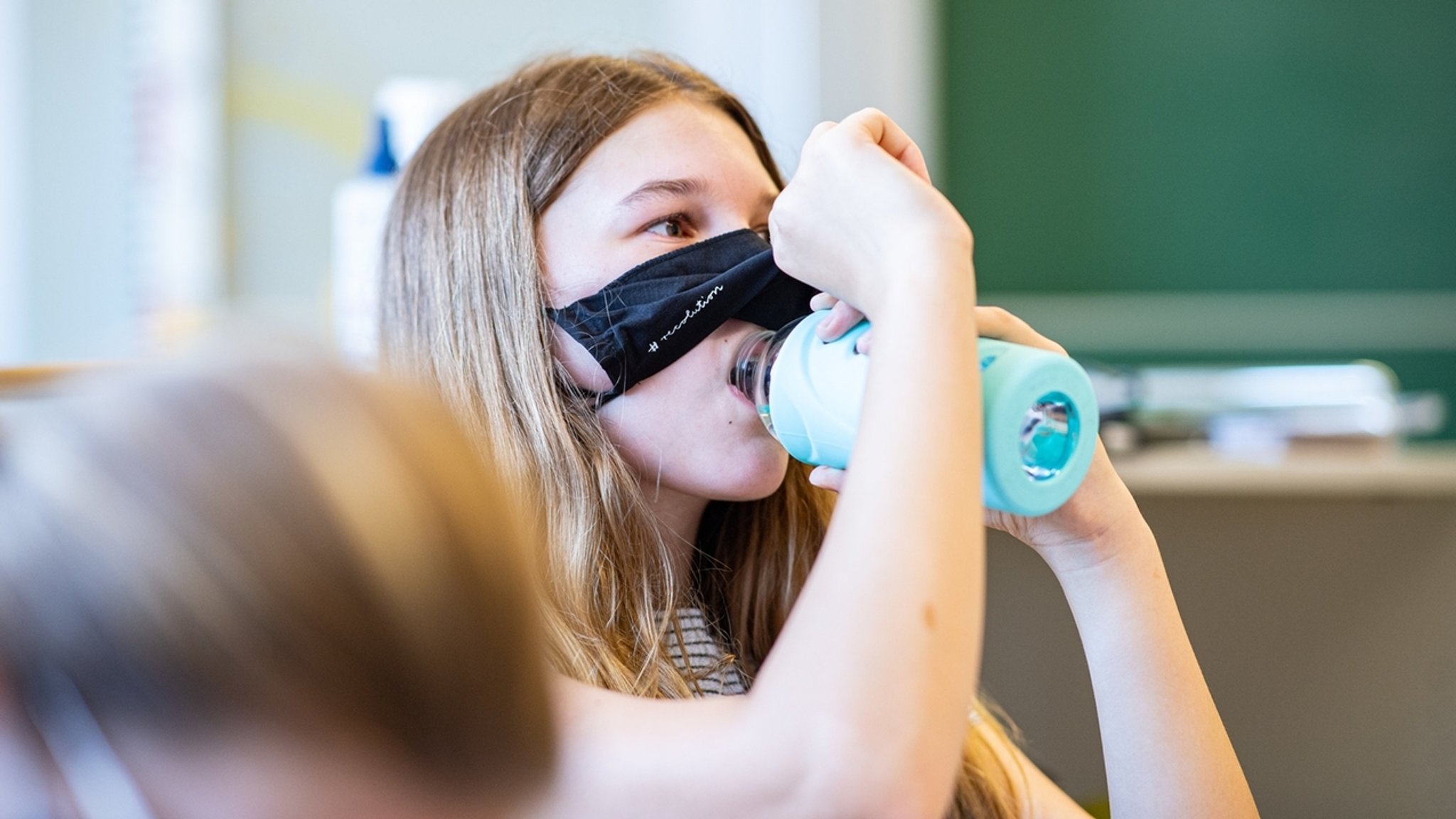 Kind schiebt die Maske hoch im Klassenzimmer, um was zu trinken.