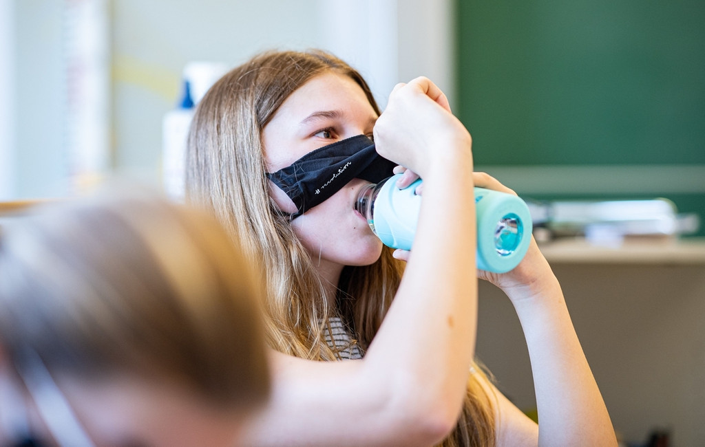 Kind schiebt die Maske hoch im Klassenzimmer, um was zu trinken.