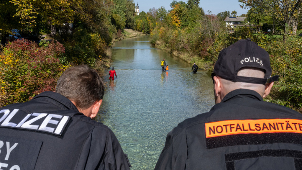 Ein Polizist und ein Notfallsanitäter sind in Rückansicht zu sehen. Sie stehen auf einer Brücke über der Prien und schauen ihren Kollegen zu, die in Neoprenanzügen den Fluss absuchen. (Archivbild)