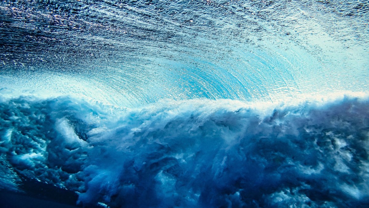 Eine Welle im Ozean, aufgenommen unter Wasser. Das Bild ist blau-weiß und zeigt in der Bildmitte eine Meereswelle.