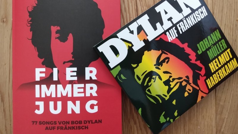Rotes Buch mit Bild des jungen Bob Dylan mit dem Titel "Fier immer jung. 77 Songs von Bob Dylan auf Fränkisch", daneben die CD "Dylan auf Fränkisch".