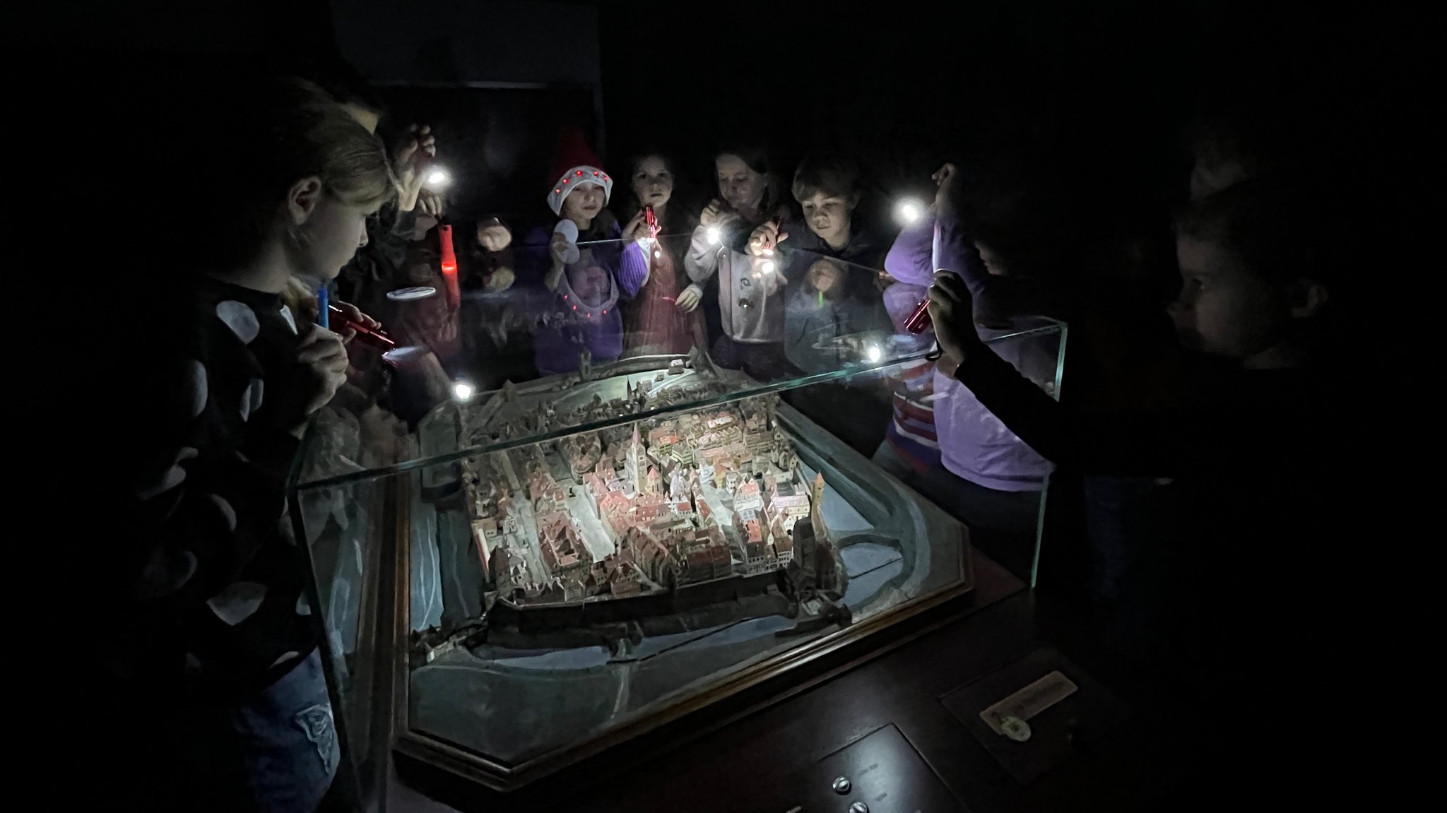 Die Kinder stehen im dunklen Museum und leuchten mit ihren Taschenlampen auf das Modell der mittelalterlichen Stadt.